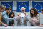 Mehrere Menschen aus verschiedenen europäischen Ländern werfen Stimmzettel in eine transparente Wahlurne vor EU-Flaggen.